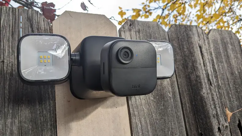 Lower front view of the Blink Outdoor 4 Floodlight Camera mounted on wooden fence outdoors with two identical floodlights located on both sides.