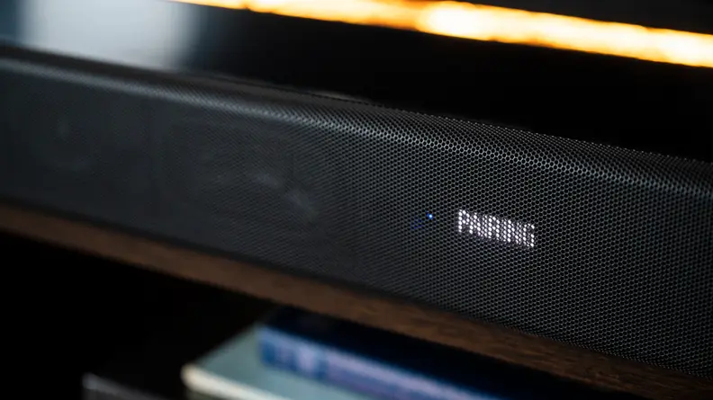 The all black Sony HT-A7000 sits atop a dark wood console against a brick wall below a large flat screen TV while the front display reads the word pairing in blue.