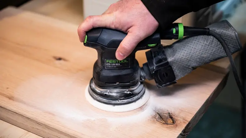 A person uses an orbital sander to sand a board