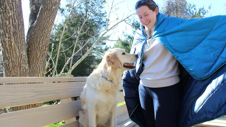 Person smiling while wearing blanketed cape on shoulders outdoors next to dog.