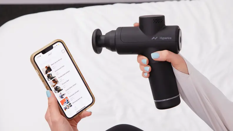 A woman holding a smartphone in her left hand and a Hyperice Hypervolt 2 Go in her right hand.