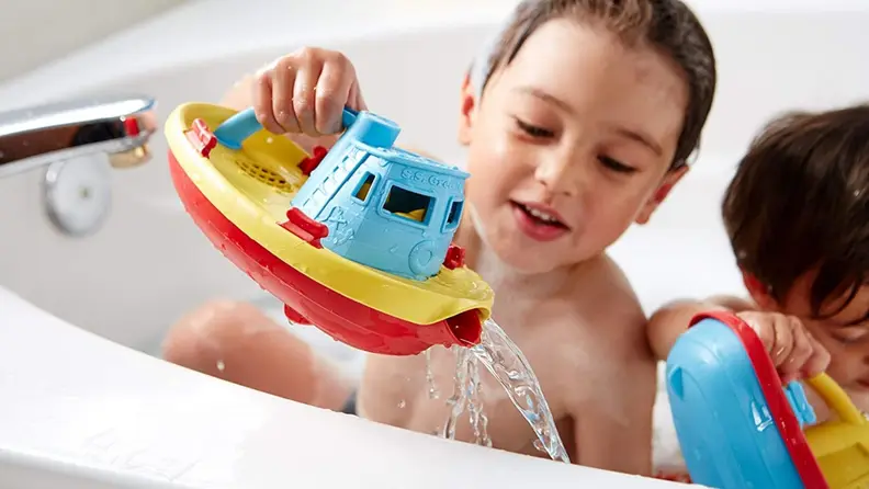A child in a bath pours water from a colorful tugboat that is made from recycled plastic.