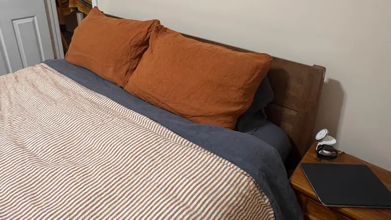 Bed made with brown and white striped comforter, navy sheets, and burnt orange pillow against FlexiSpot Kana brown headboard, in front of a white wall.