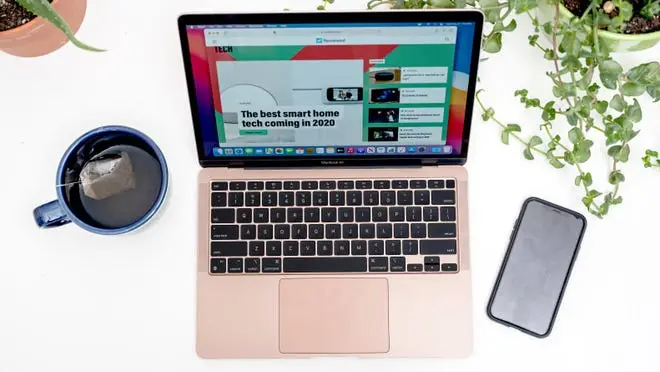 Apple laptop on desk next to smartphone and hot cup of tea.