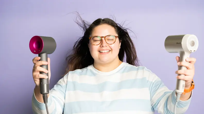 A person wearing a blue and white striped shirt standing in front of a purple background and pointing to hair dryers at their long brown hair while smiling.