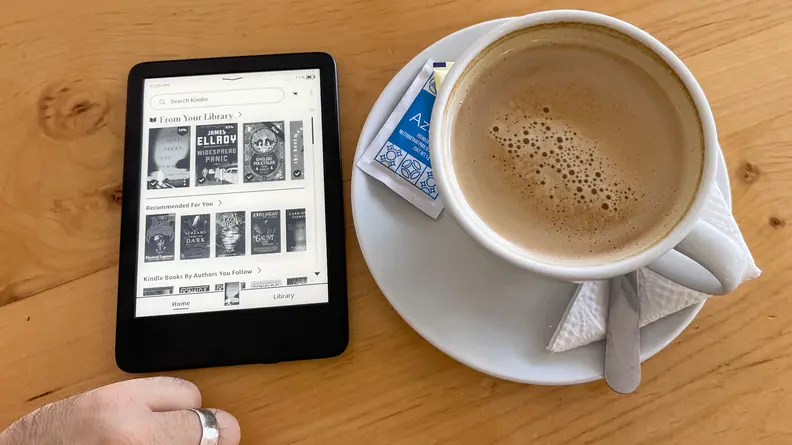 An Amazon Kindle e-reader sits on a table next to a large cup of coffee
