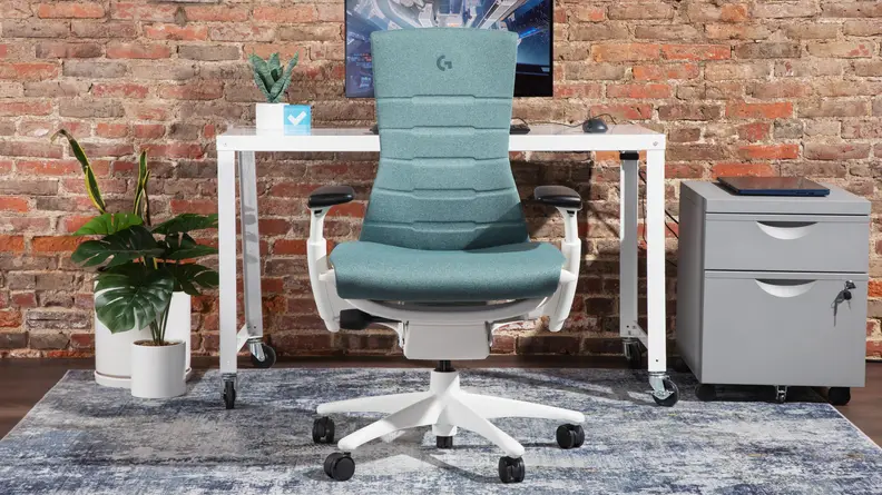 The Herman Miller x Logitech G Embody gaming chair in an office space, next to a white desk, computer monitor, and office supplies.