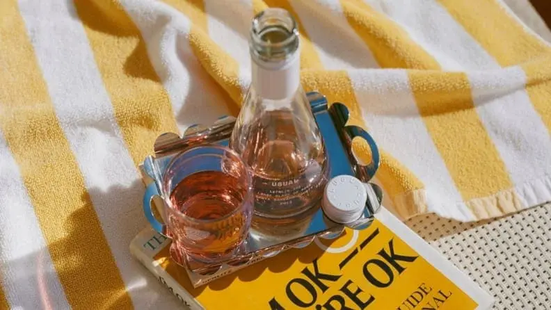 A small glass of wine is sitting next to a small wine bottle atop a white and yellow striped towel with a book nestled underneath. It's a very sunny, almost beach-like scene.