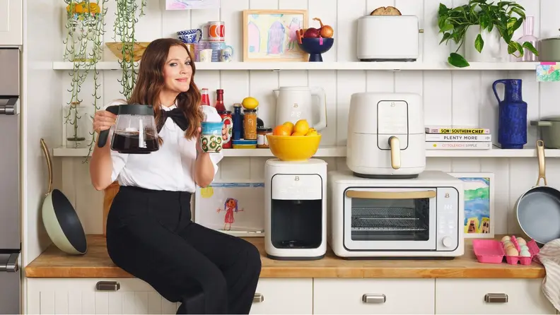 Drew Barrymore sitting on her kitchen counter surrounded by her appliance line.