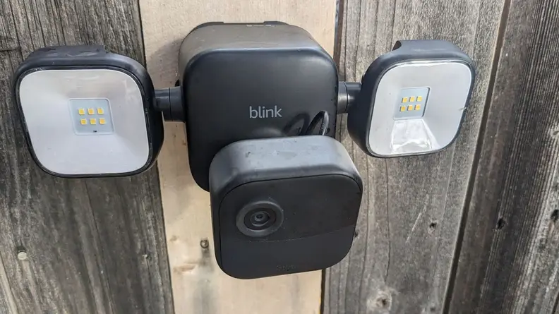 Downward aerial view of the Blink Outdoor 4 Floodlight Camera mounted on wooden fence outdoors with two identical floodlights located on both sides.