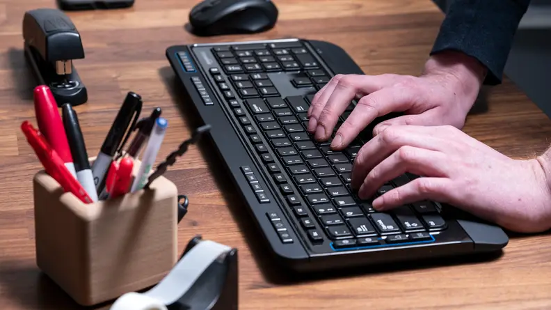 A black wireless keyboard sits on a wooden desktop; a wireless mouse, a stapler, a pen holder, and a tape dispenser surround it. Two hands are typing on the keyboard.
