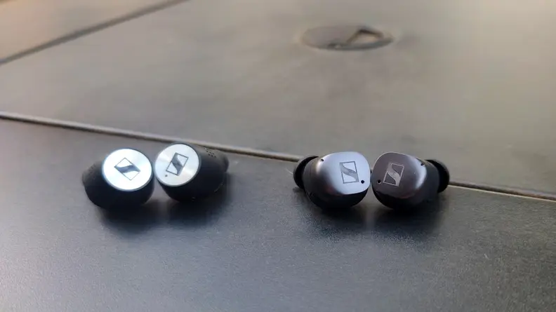 The silver capped Sennheiser Momentum True Wireless 2 set next to the graphite Momentum True Wireless 3 on a black table.