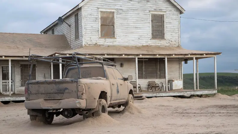 A pickup truck partially buried in sand outside a dilapidated farmhouse.