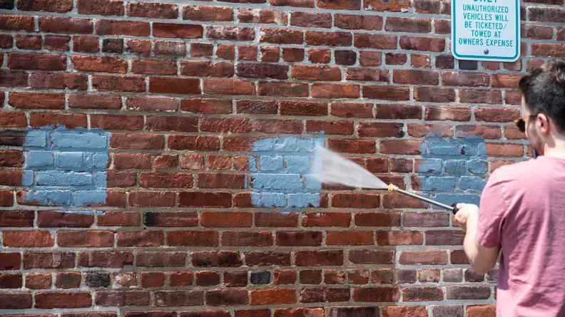 A person sprays a blue painted square on a brick wall with a pressure washer.