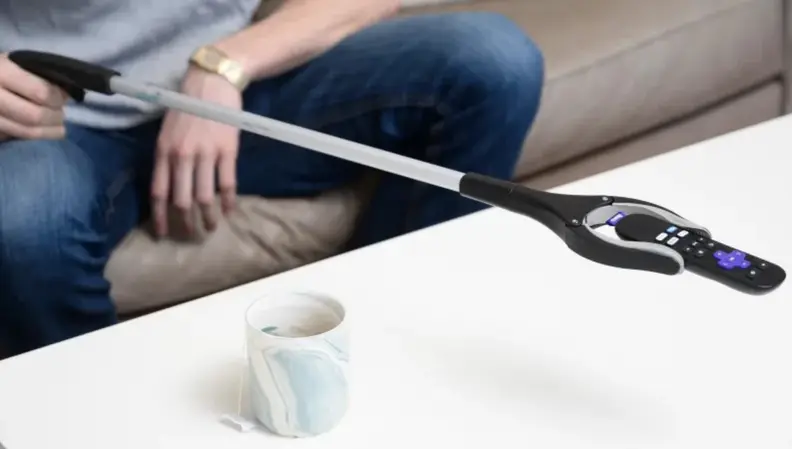 A person holds a remote control using the Vive Rotating Reacher Grabber Tool over a white coffee table and coffee mug.