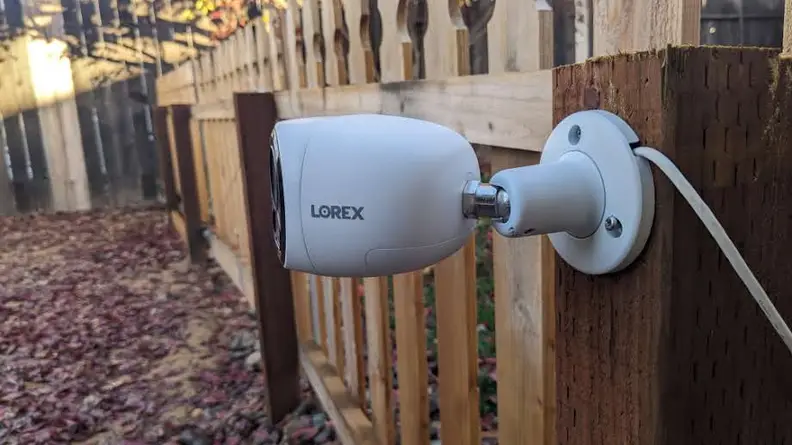 A side angle view of the Lorex 4K Dual-Lens Wi-Fi Security Camera hanging on a fence outdoors.