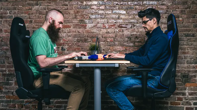 Two testers sitting opposite each other in some of the best gaming chairs on the market right now, against a brick wall