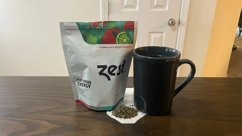 A bag od Zest Pomegranate Mojito Green tea sits on a table next to a  mug. Loose tea leaves sit under the mug on a paper towel.