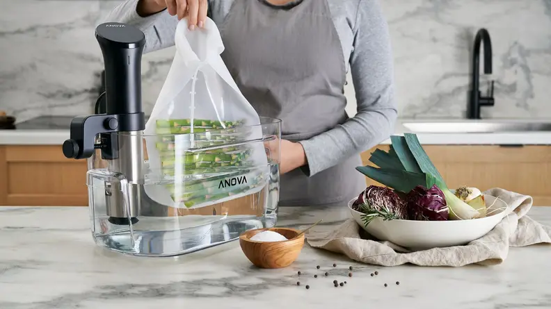 In a spacious kitchen, a person is lifting a bag of asparagus cooked by an Anova immersion circulator. A bowl of uncut leaks and purple vegetables and some salt are next to the person.