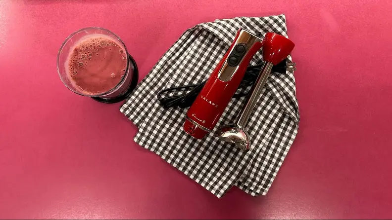 The Galanz Retro Immersion Hand Blender in the color red, laying on a black and white checkered towel, next to a glass of red juice.