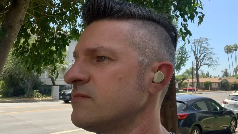 A man stands outside on a sidewalk looking off camera while wearing the gold beige Jabra Elite 5 earbuds.