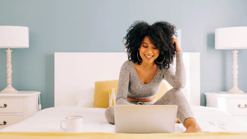 A woman sits comfortably on her bed in cozy loungewear, smiling while working on her laptop, showcasing the convenience of fast and reliable T Mobile 5G Home Internet.
