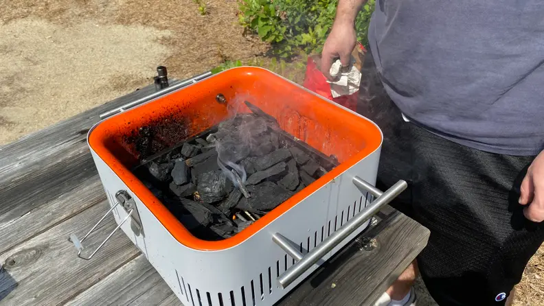 A person is standing beside an Everdure Cube grill in gray, which is packed with burning charcoal.