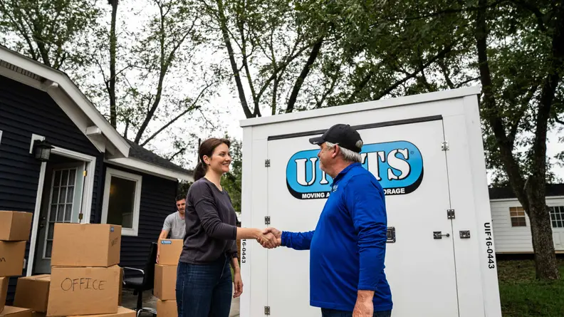 Two people talk in front of a portable storage unit surrounded by moving boxes