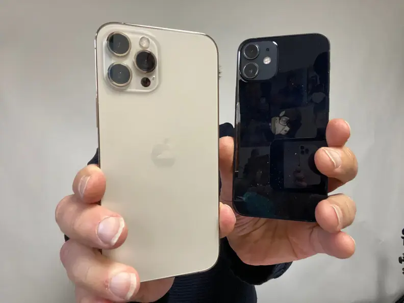 A person holding two Apple iPhone models. In the left hand a white 12 Pro Max and the right, a black 12 Mini.