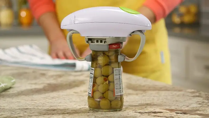 A hands-free electric jar opener opens a jar of olives on a marble countertop with a person in a yellow shirt standing in the background.