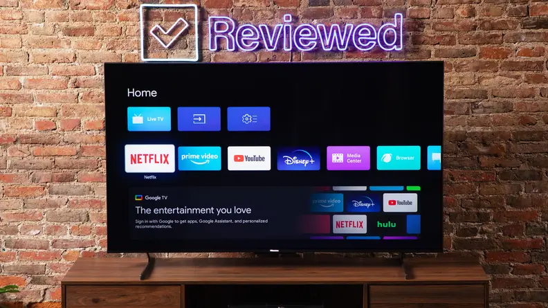 The Hisense U8H on a wooden credenza displaying Google OS with a brick wall in the background and a Reviewed neon sign.