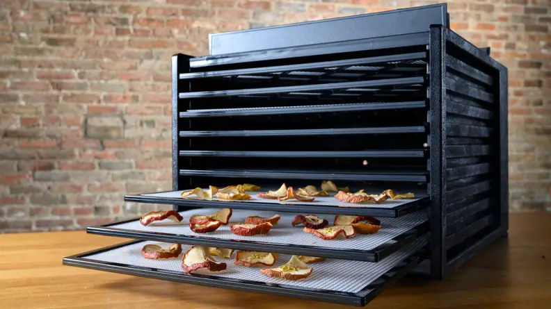 The Excalibur Food Dehydrator perched on a wooden table with a few of its bottom trays pulled out to reveal dehydrated apple segments.