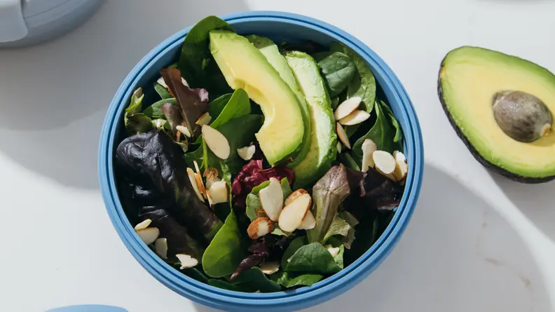 In the center of the image, there's a Stojo bowl filled with salad with sliced avocado and almonds on top. A half avocado is next to it.