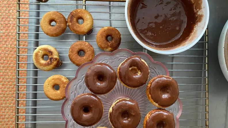 Unglazed mini donuts on a drying rack next to a pink glass tray filled with chocolate glazed mini donuts.