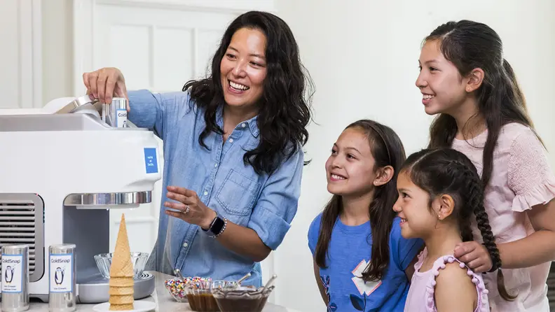 A parent and several children gather, smiling, around a large white machine. Ice-cream cones and toppings are on the table beside it.