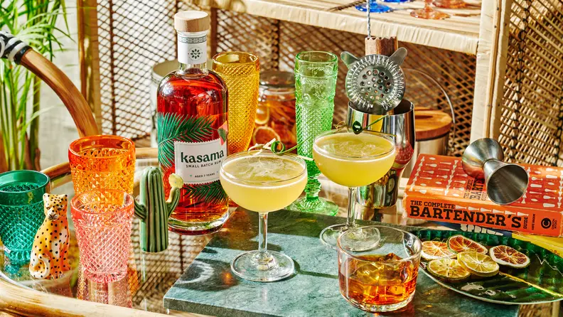 On a cocktail table, a bottle of Kasama aged rum is the main focus. Around the bottle there are a few glasses of cocktails.
