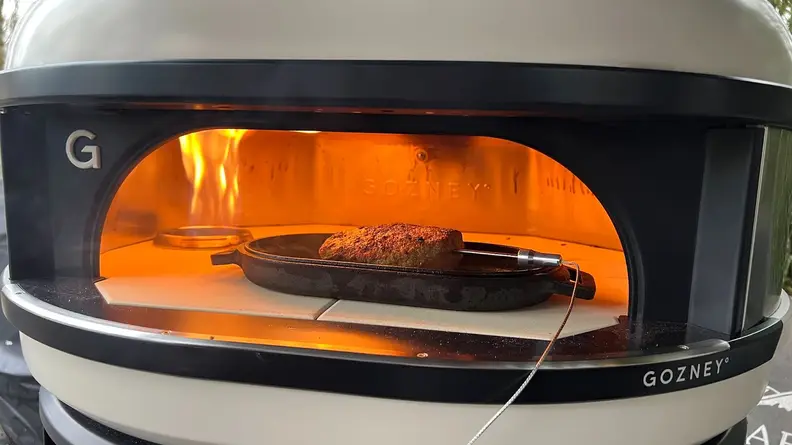 Meat cooking with temperature probe inserted in roast, inside of the Gozney Dome Pizza Oven next to open flame.