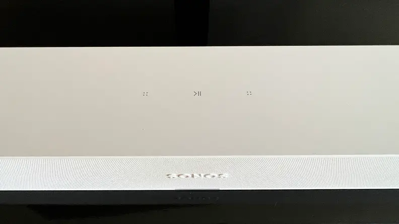 The white Ray soundbar's top touch keys are shown as it sits on a black and brown console.