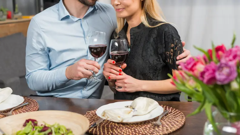 A romantic dinner setting featuring a couple dressed elegantly, toasting with glasses of red wine. The table is set with white plates, woven placemats, and cloth napkins, with a fresh salad in the foreground and vibrant pink flowers adding a romantic touch. The couple leans in close, smiling, creating a cozy and intimate atmosphere.