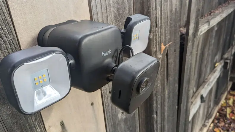 Side view of the Blink Outdoor 4 Floodlight Camera mounted on wooden fence outdoors with two identical floodlights located on both sides.