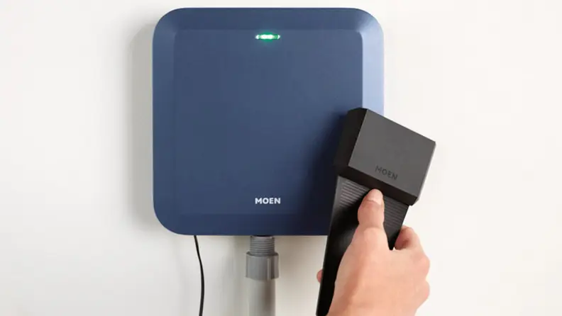 Person holding black soil sensor in front of Moen Smart Sprinkler Controller that’s mounted on a wall.