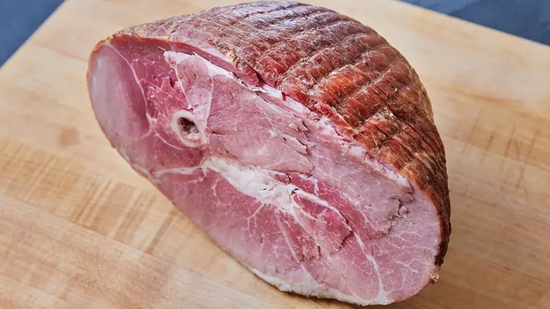 A bone-in honey baked ham sits on a light wood cutting board.