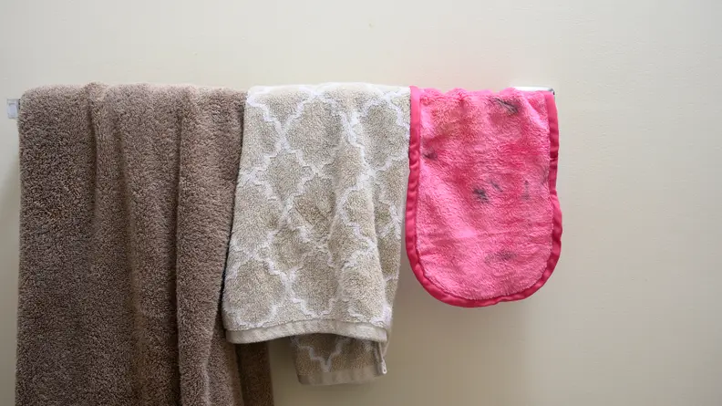 The Makeup Eraser hung over a towel ring next to a hand towel and bath towel.