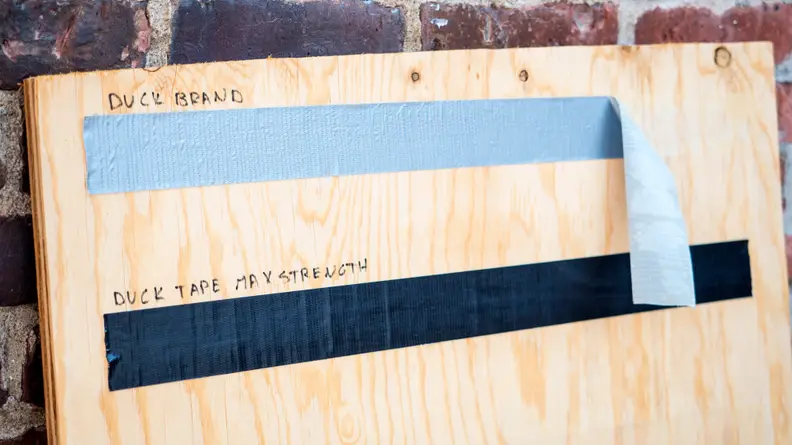 Duct tape strips on plywood after 4 weeks. The upper Duck brand tape has started to peel. The Duck Tape Bax Strength below it is holding fast.