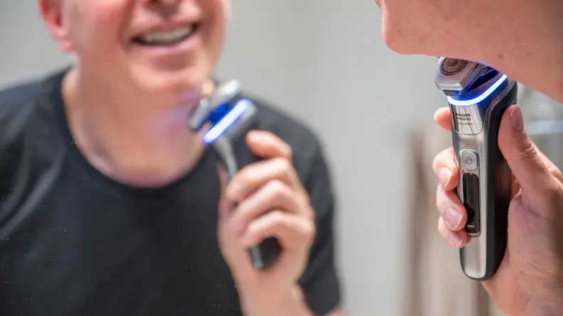 A man looks in his bathroom mirror as he shaves with an electric razor.