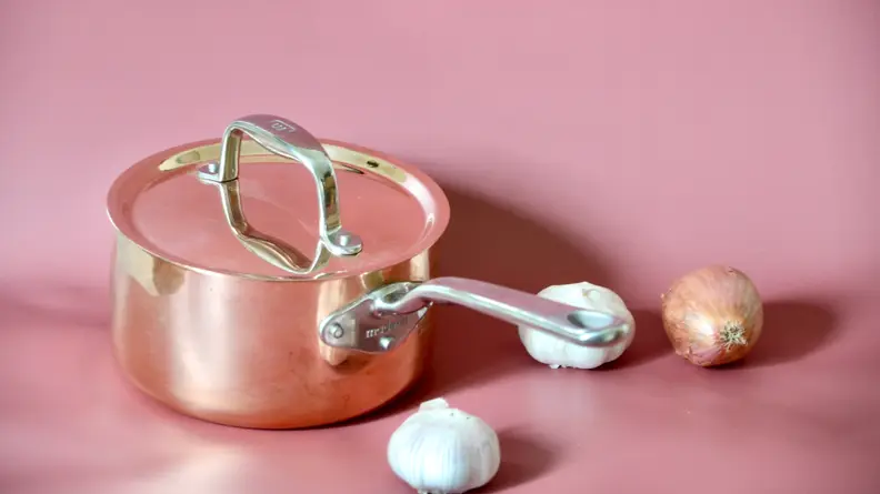 A copper pot with a lid on a pink background next two a garlic bulb and an onion.