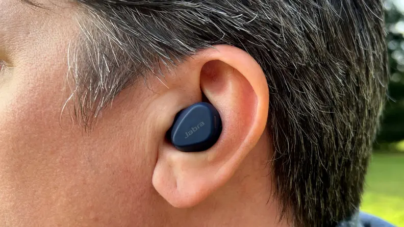 A navy earbud sits in the ear of a man with brown and grey hair with trees and grass in the background.
