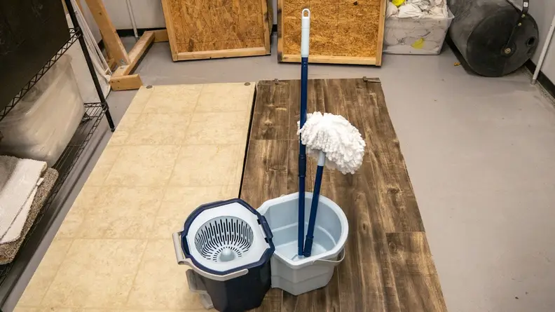 The Casabella Clean Water Spin Mop sits in a room prepped for testing, with both hardwood flooring and linoleum sections set up beneath it; the mop handle is disassembled and propped up in the water bucket.