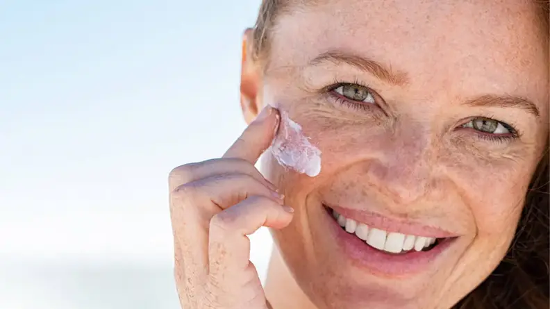 A person smiling at the camera while applying a white cream to their face.