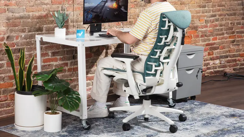 The side view of the Herman Miller x Logitech G Embody near a white desk, with a computer monitor and office supplies.